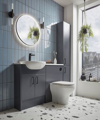 Stylish modern bathroom featuring glossy blue vertical tiles, a sleek vanity unit with integrated sink, and matte charcoal cabinetry for smart storage. A round backlit mirror, hanging plants, and terrazzo flooring.