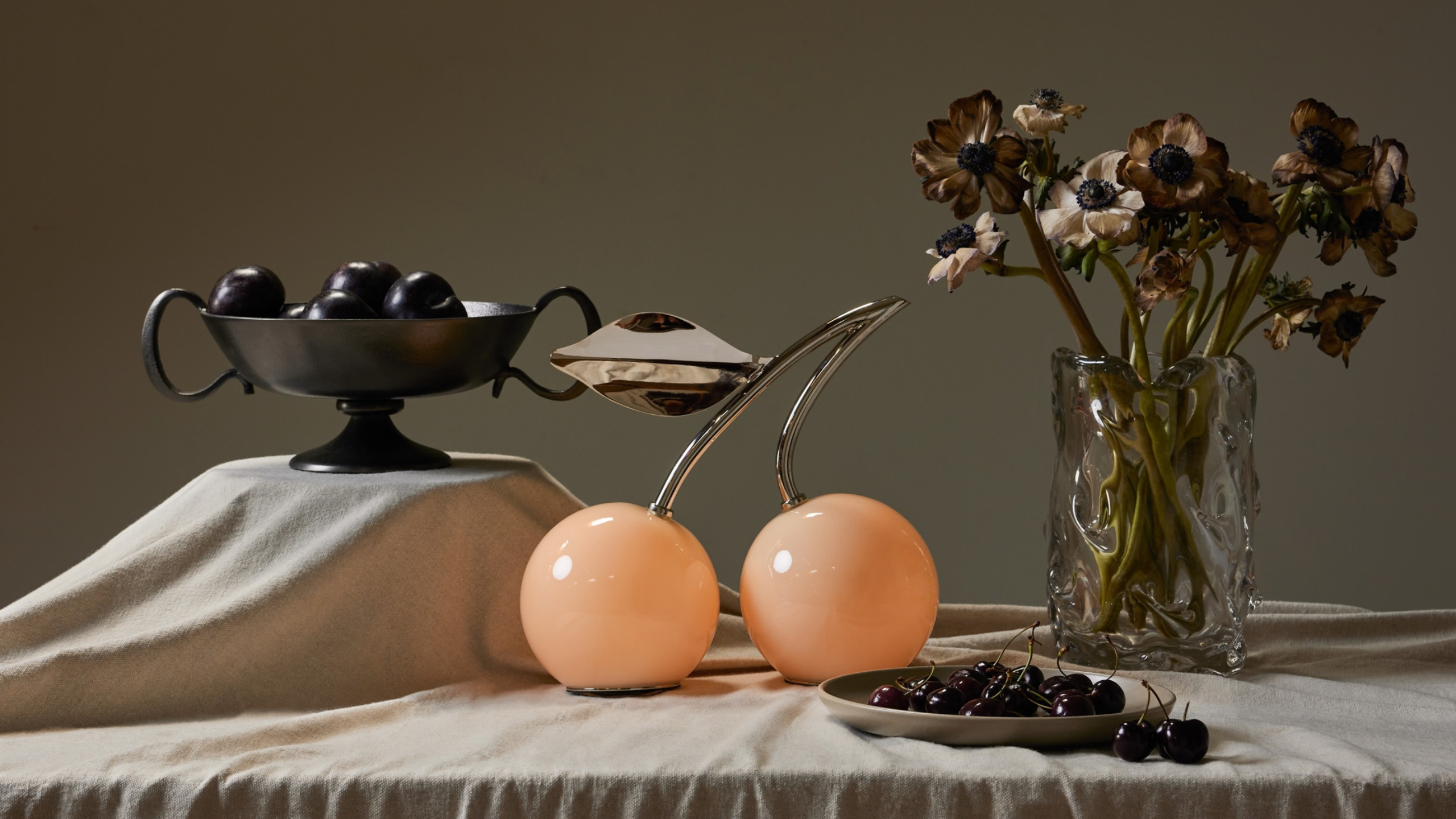 Minimalist tablescape with draped linen, a sculptural fruit bowl, an irregular vase of flowers, and a cherry-inspired table light. 