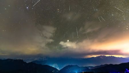 Shooting stars are pictured streaking downwards through a starry sky towards a cloudy horizon lined with mountains, lit in a blue mist.