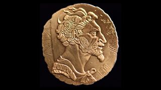 Attila the Hun&#039;s face on a gold-looking coin with his name around the edge. His face is facing right and he has lots of tuffs of short, wild hair, large ears, a strong brow, large nose, and a beard and moustache. 