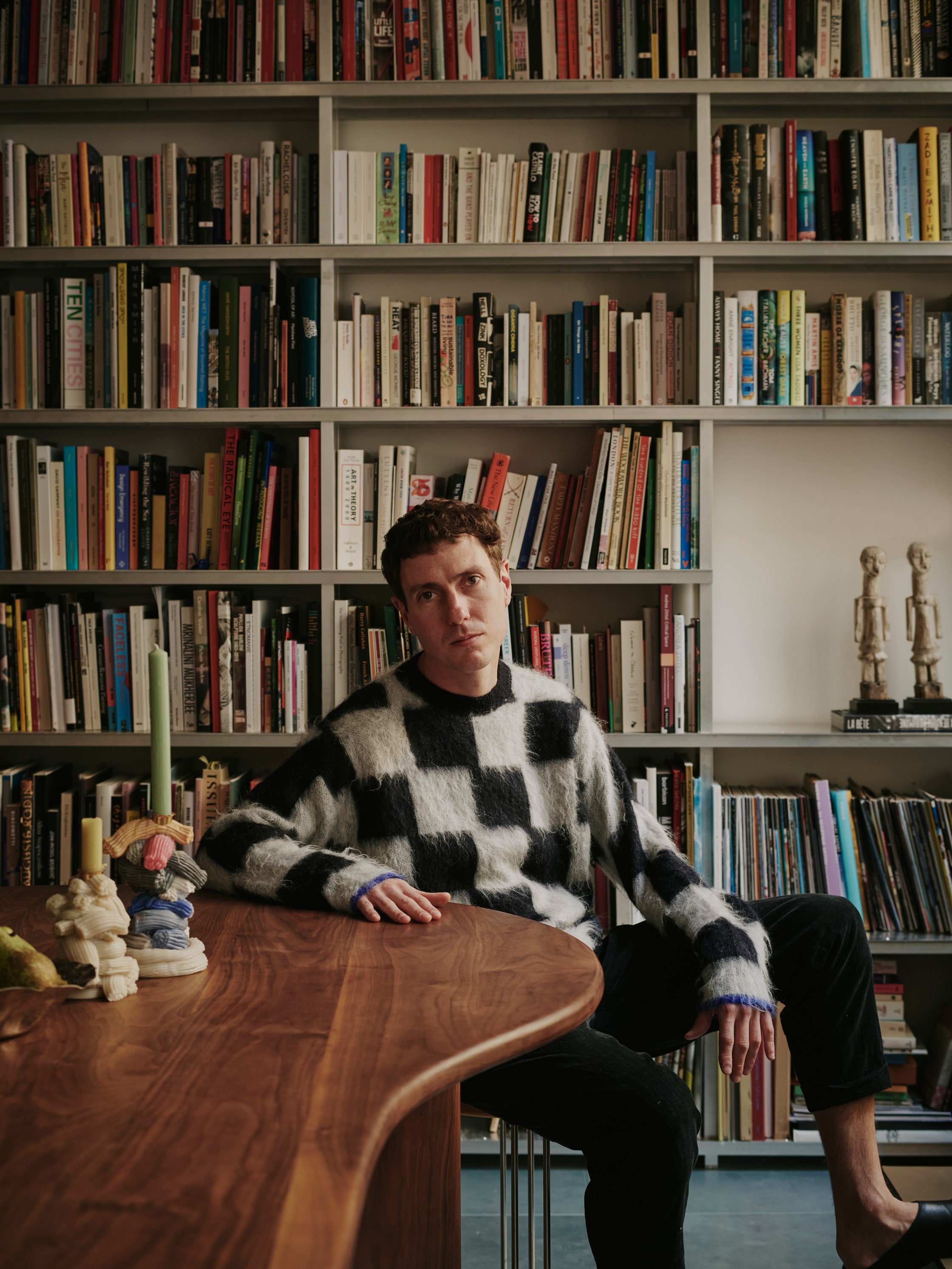 A young man dressed in a checkered black and white jumper, black trousers, and leather black pumps sits at a wavy wooden table next to squishy candelabra in blue, cream, pink, and yellow tones, in front of a stacked-with-tomes bookshelf.