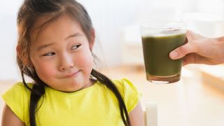 A child scrunches up her face at a green smoothie being held up beside her