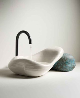 Daniel Arsham Kohler sink made of 3D printed white ceramic resting on an oxidised brass stone-shaped object, with water coming out of a minimalist black tap