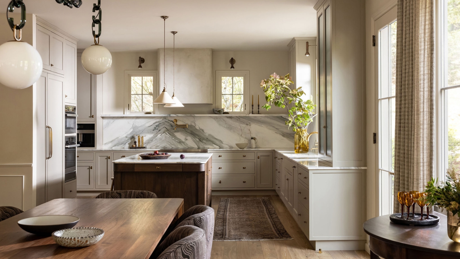 Kitchen with pale gray cabinets, a walnut island, veined marble backsplash and cream pendant lights, beside a wood dining table.
