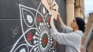 An artist wears a grey hoodie and head covering while using a cardboard stencil to spray-paint an intricate, circular geometric mural onto a black brick wall.
