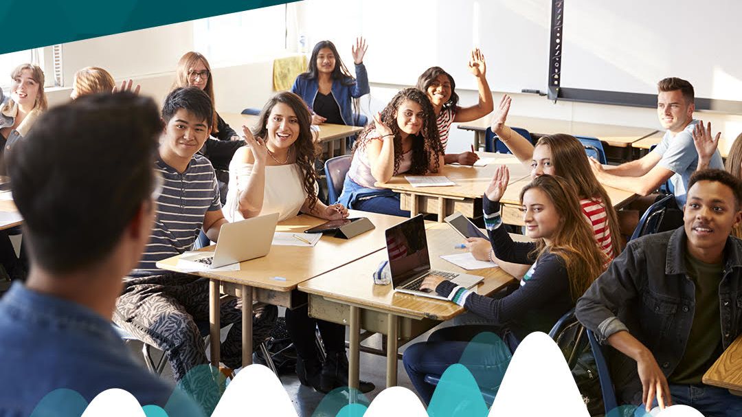 Smiling students using laptops raise their hands in a classroom.