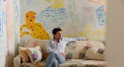 A young man dressed in a light blue shirt and denim jeans sits on a beige couch with colorful illustrations in front of a murals-covered wall depicting monsters and fantastical creatures.