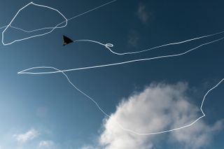 Daytime image, white squiggly digital line attached to a grey inflated aeroglyphic, representing the trail when in flight, blue sky fluffy white clouds
