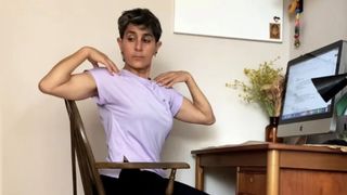 Fitness editor Yanar Alkayat is sat in a chair at her desk performing spine rotations. Her fingertips rest on her shoulders and her elbows are out to the sides as she twists her torso to the right. In front of her is a Mac desktop computer and dried flowers in a case.