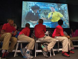 four students on chairs twist around to take a look at a screen behind them. the screen has two astronauts floating on the international space station, side by side. the astronaut on the right holds a microphone