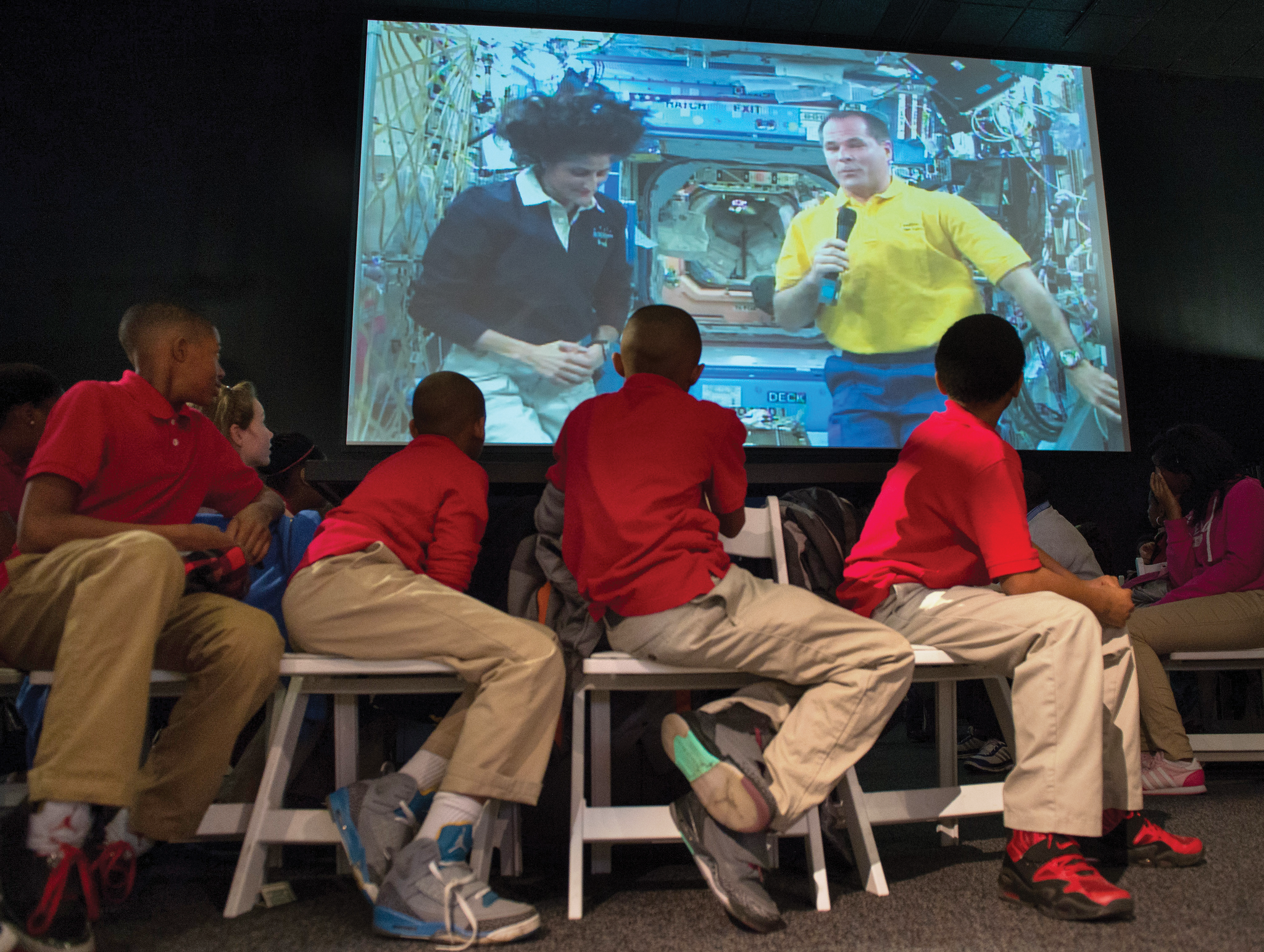 Quatre étudiants se recroquevillent sur des chaises pour regarder un écran derrière eux.  Sur l'écran, deux astronautes flottent côte à côte sur la Station spatiale internationale.  L'astronaute de droite tient un microphone