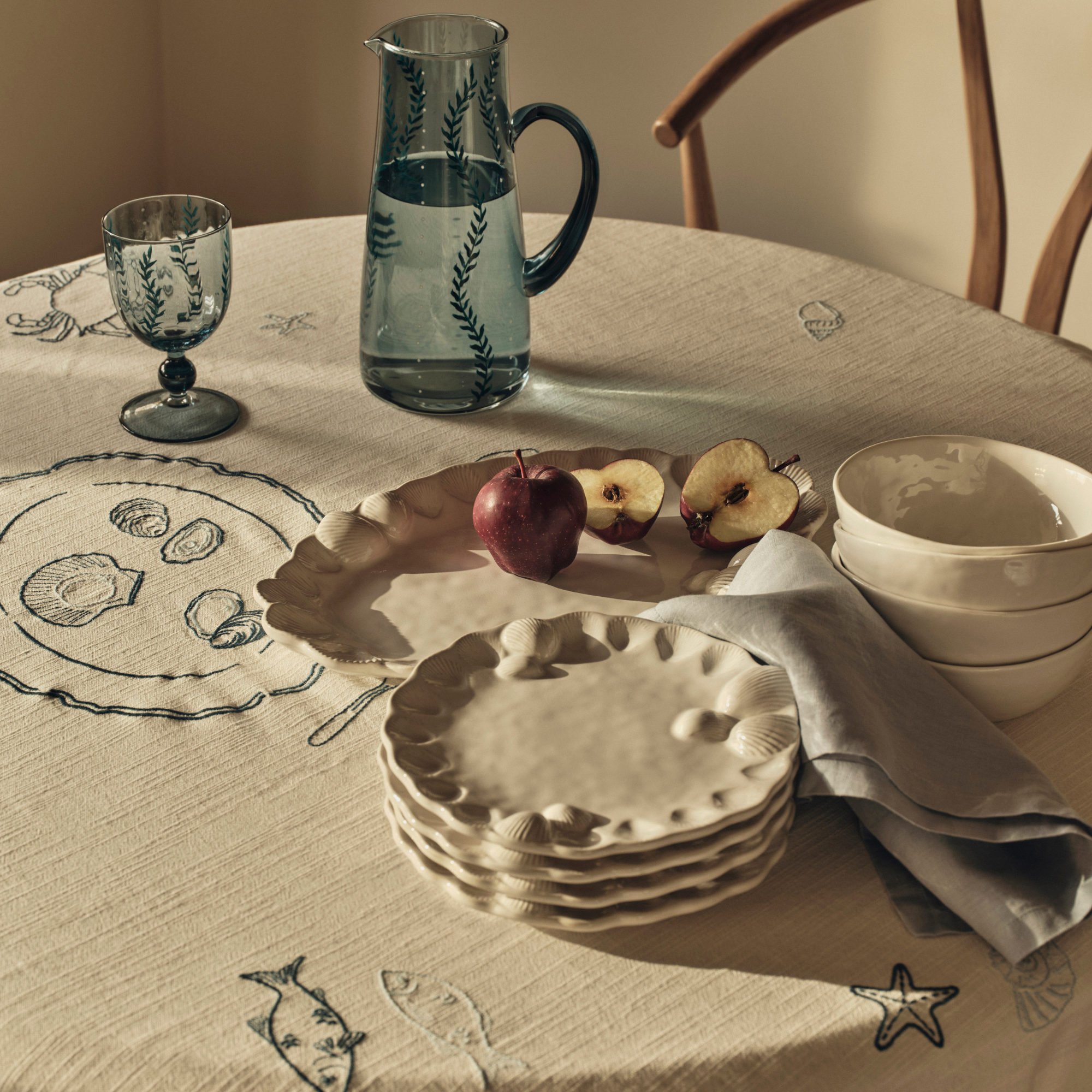 Pure Cotton Coastal Embroidered Tablecloth, white plates, blue jug and wine glass, and cut apples on circular table.