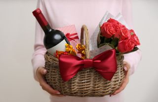 A person holding a wicker hamper full of gifts including red roses, and a bottle of wine.
