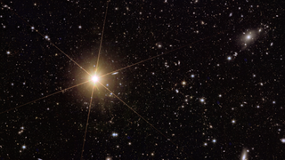 The dark background of space with an incredibly bright galaxy in the middle-left of the image. It has diffraction spikes thanks to the telescope's instruments and there are lots of other bright, albeit way less bright, speckles throughout the image.