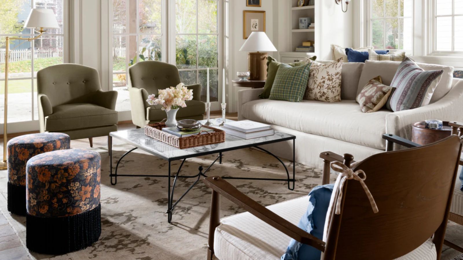 a living room with a large beige couch with pillows on top, two green accent chairs, a large coffee table with decor on top, and two floral ottomans 