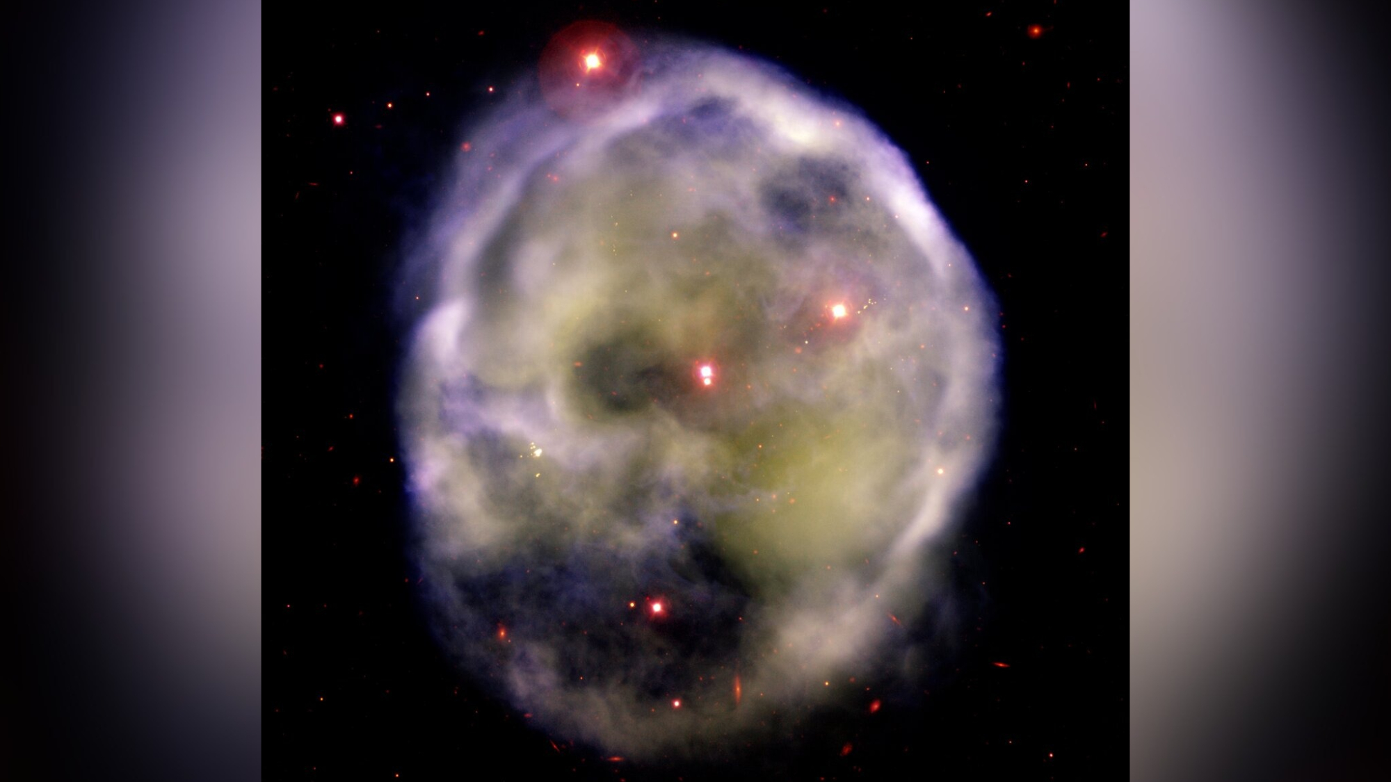 A glowing, roughly circular nebula with wispy blue-green gas and reddish patches against a dark, star-filled background, resembling a ghostly skull.