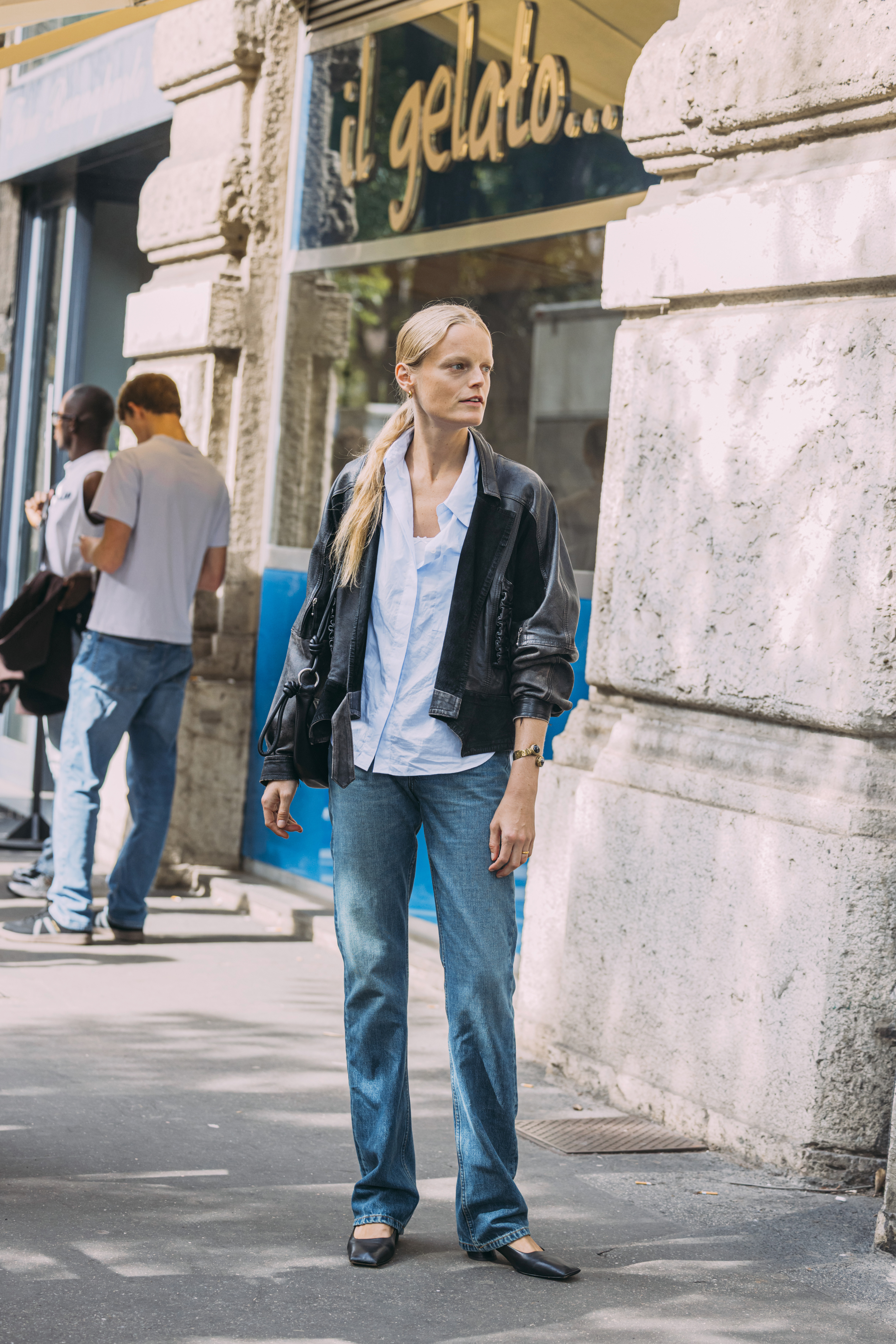 A woman wears a leather jacket, blue button-down shirt, stovepipe jeans, and black pumps.