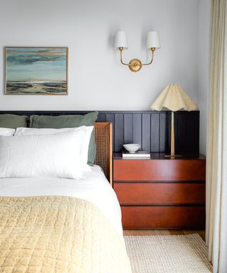 White bedroom with dark blue paneling, bed with white bedding, dark wood bedside cabinet, table lamp, wall light, artwork above bed