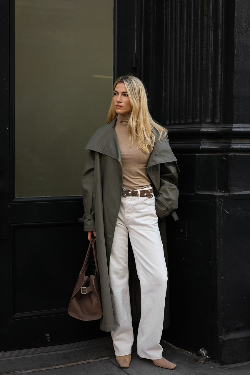 How to style white jeans for fall, shown in a photo of a woman standing outside against a black building wearing a green trench coat over a tan turtleneck with a tan studded belt, white wide-leg jeans, tan suede ankle boots, and a brown bowler bag