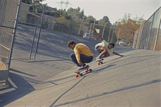 Daytime image of Chuck Askerneese and Marty Grimes at Kenter Canyon, 1975, skateboarding crouched on a concrete slop, metal mesh fences, trees and buildings in the distance