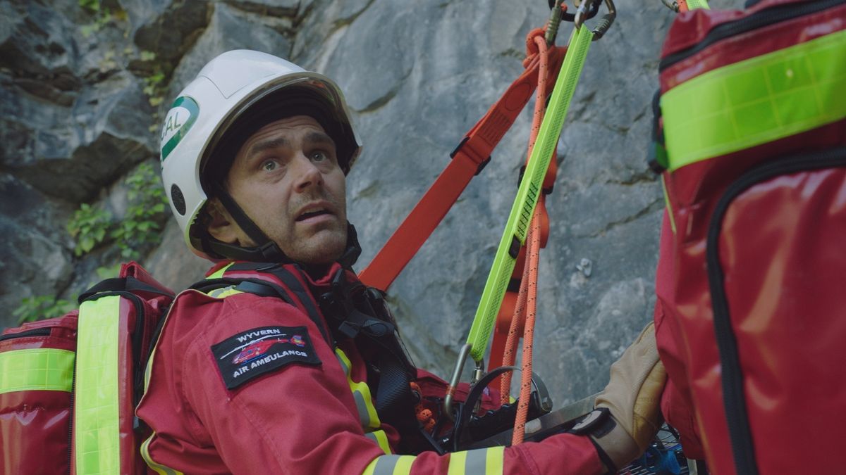 Iain Dean strapped up in red HEMS uniform on a cliff face in Casualty episode Precipice. 