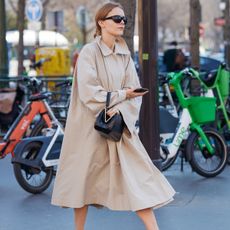 a woman wears a long beige trench dress with a black bag.