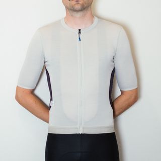 A white man in a taupe cycling jersey and black cycling shorts stands against a white wall