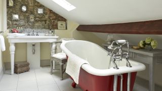Red free-standing bath with stone wall and decorative mirrors, antique chairs and white tiled floor