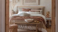 A neutral bedroom with a fluffy, layered bed, viewed through a door frame. A wooden bench at the base of the bed with a large white bowl on it. 