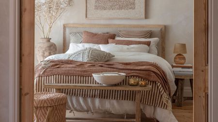 A neutral bedroom with a fluffy, layered bed, viewed through a door frame. A wooden bench at the base of the bed with a large white bowl on it. 