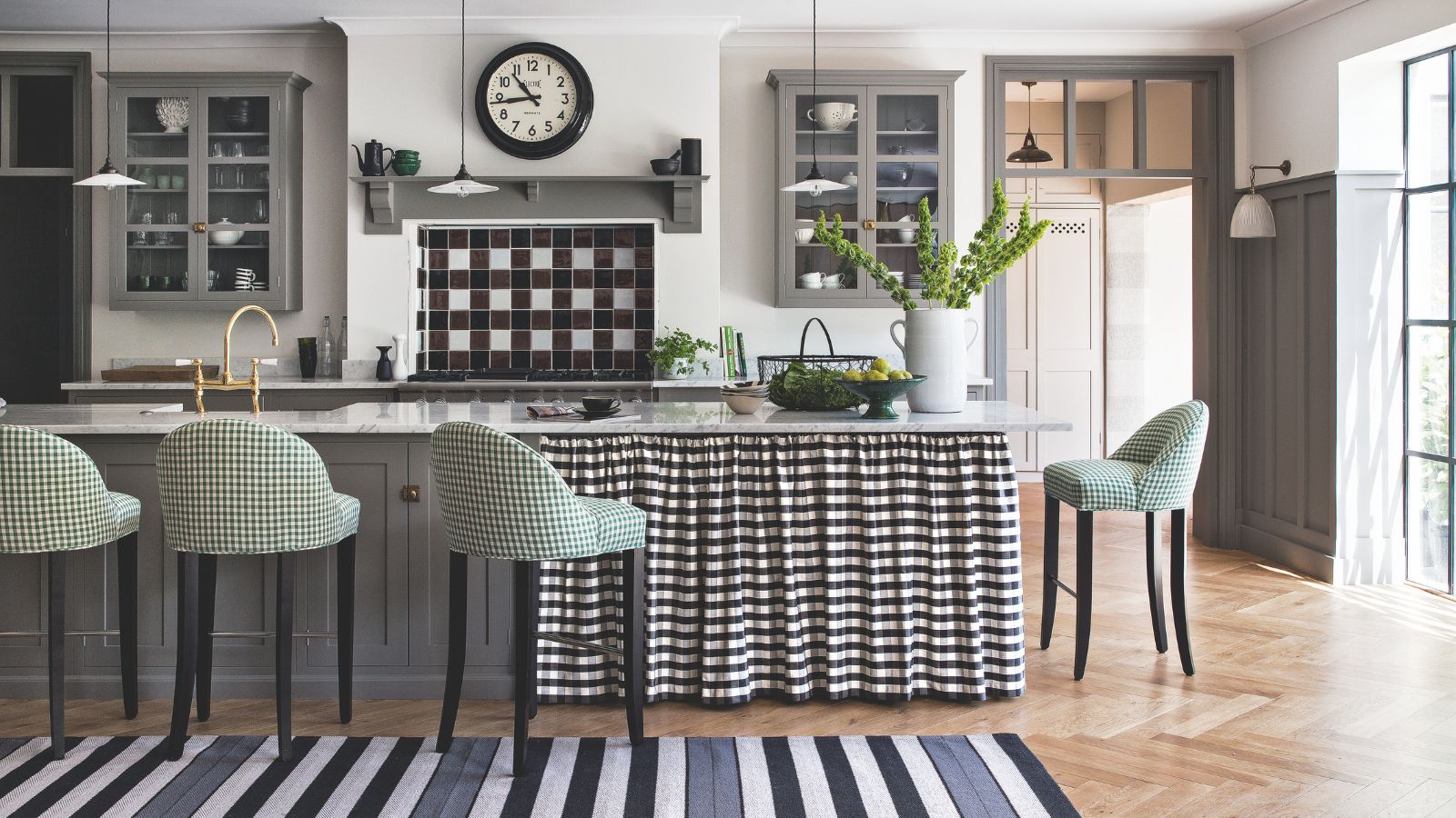 A black and white kitchen with grey cabinets and a wooden floor. In front of the black and white tiled hob is a marble island with a black and white gingham curtain, surrounded by green and white stools. In the foreground is a black, grey and white striped rug. Above the hob is a black clock, and to the right is an open doorway to a bright white room behind. There is a bright window to the far right.