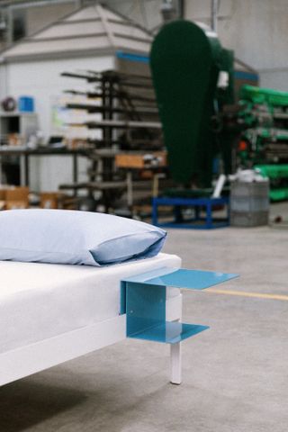 The bed with white frame, featuring a blue side table and light blue pillow. Photographed inside the factory space