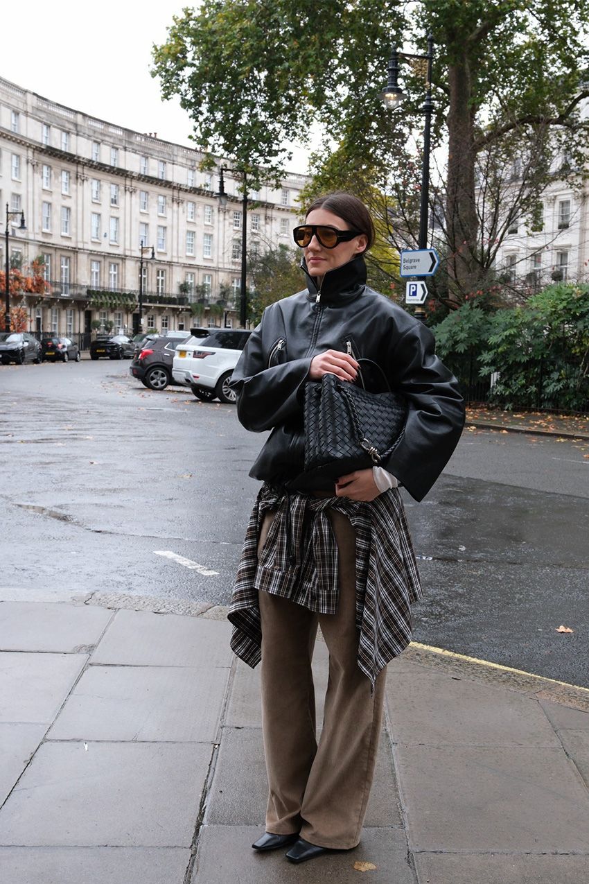A woman standing outside on the sidewalk wearing a black leather funnel neck bomber jacket styled with brown wide-leg pants, a brown plaid flannel tied around the waist, black ankle boots, a black handbag, and two-tone aviator sunglasses.