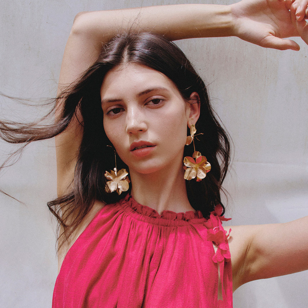 Young brunette woman looking at camera, wearing Fabrique pink dress and big gold flower earrings.