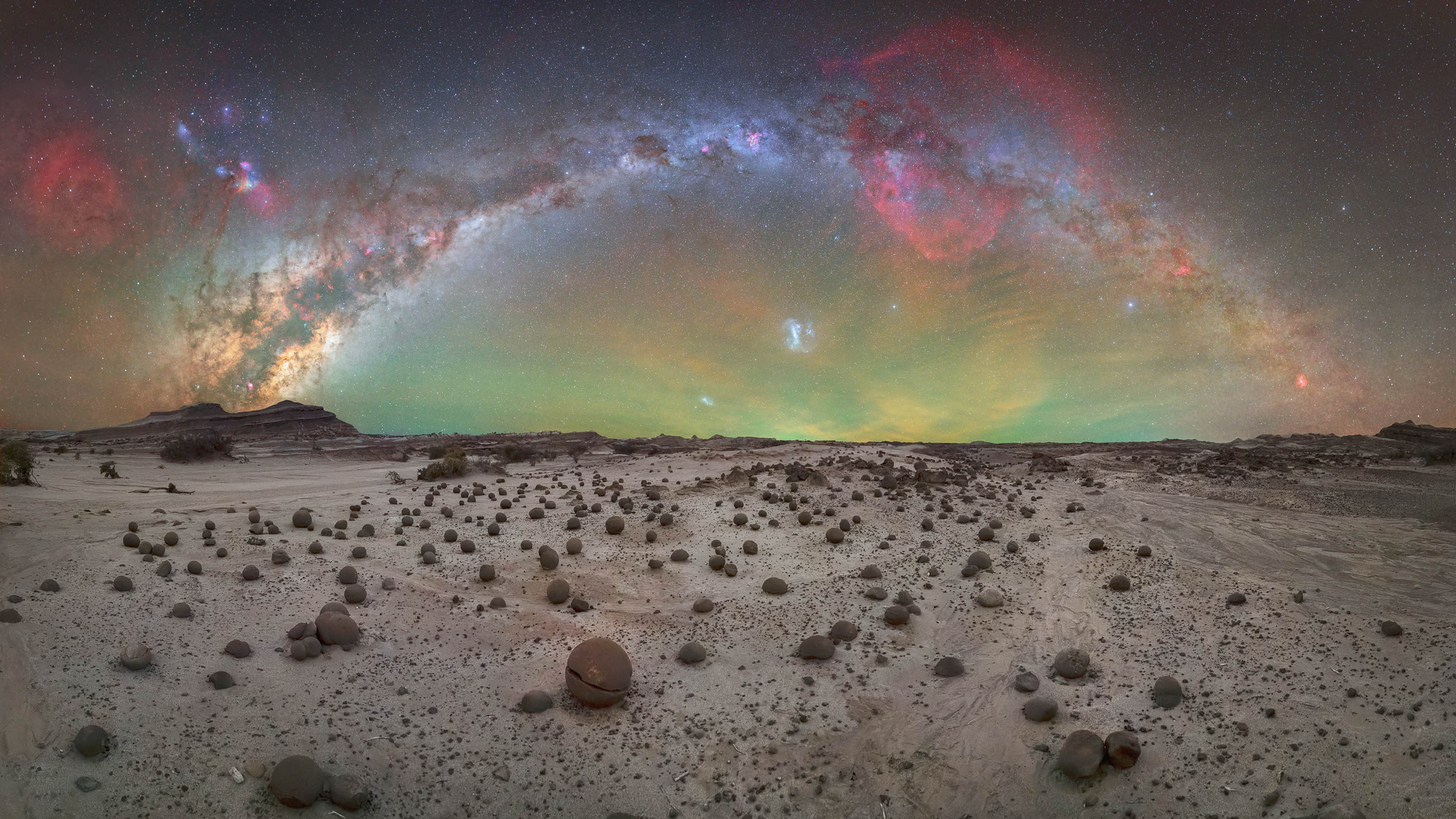 Desert landscape scattered with round stones under a vivid Milky Way arch and colorful night sky