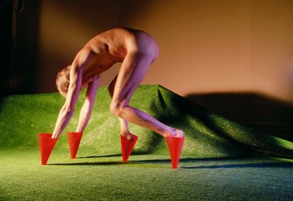 Naked white male model, arched over with hands and feet in small red cones, on a grass effect green carpet, brown wall backdrop
