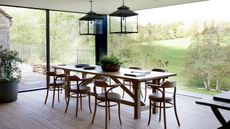 Large dining room with floor-to-ceiling windows revealing open green space. Inside is a wooden dining table, wooden chairs, wooden floor two lampshades and a potted plant.