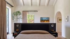 A large, bright, off-white bedroom with windows and doors around the room that have tan blackout curtains on rods. There is a bed in the middle of the room with a black headboard and beige bedding.