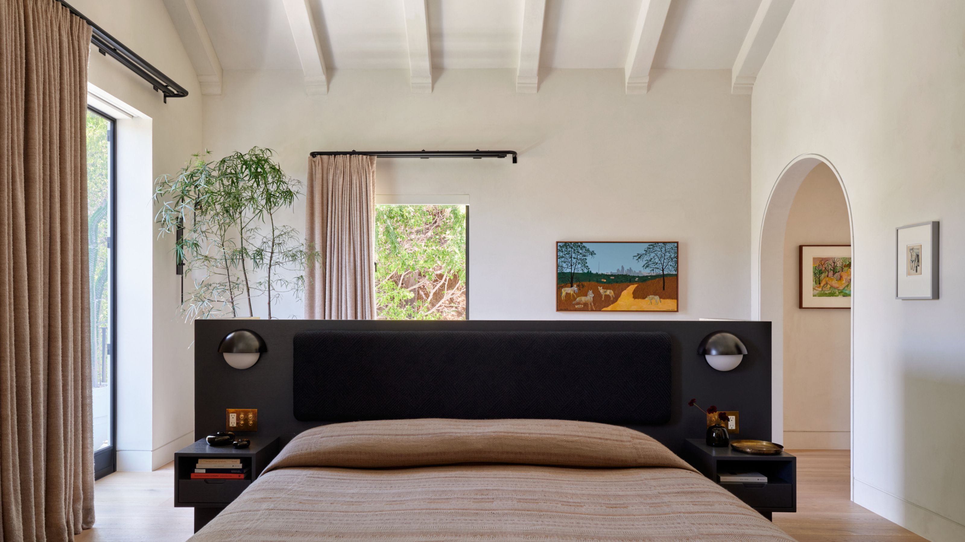 A large, bright, off-white bedroom with windows and doors around the room that have tan blackout curtains on rods. There is a bed in the middle of the room with a black headboard and beige bedding.