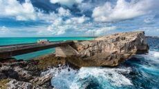 A van crosses a bridge, with turquoise water one one side and waves breaking on rocks on the other, at the Glass Window bridge, Eleuthera island, Bahamas