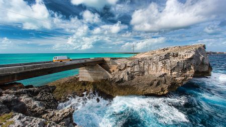 A van crosses a bridge, with turquoise water one one side and waves breaking on rocks on the other, at the Glass Window bridge, Eleuthera island, Bahamas