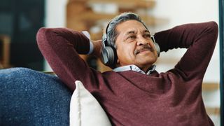 an older man with brown skin and a salt-and-peppered mustache reclines on a sofa with overear headphones on. His hands are behind his head, as if he's resting, and he's looking into the distance