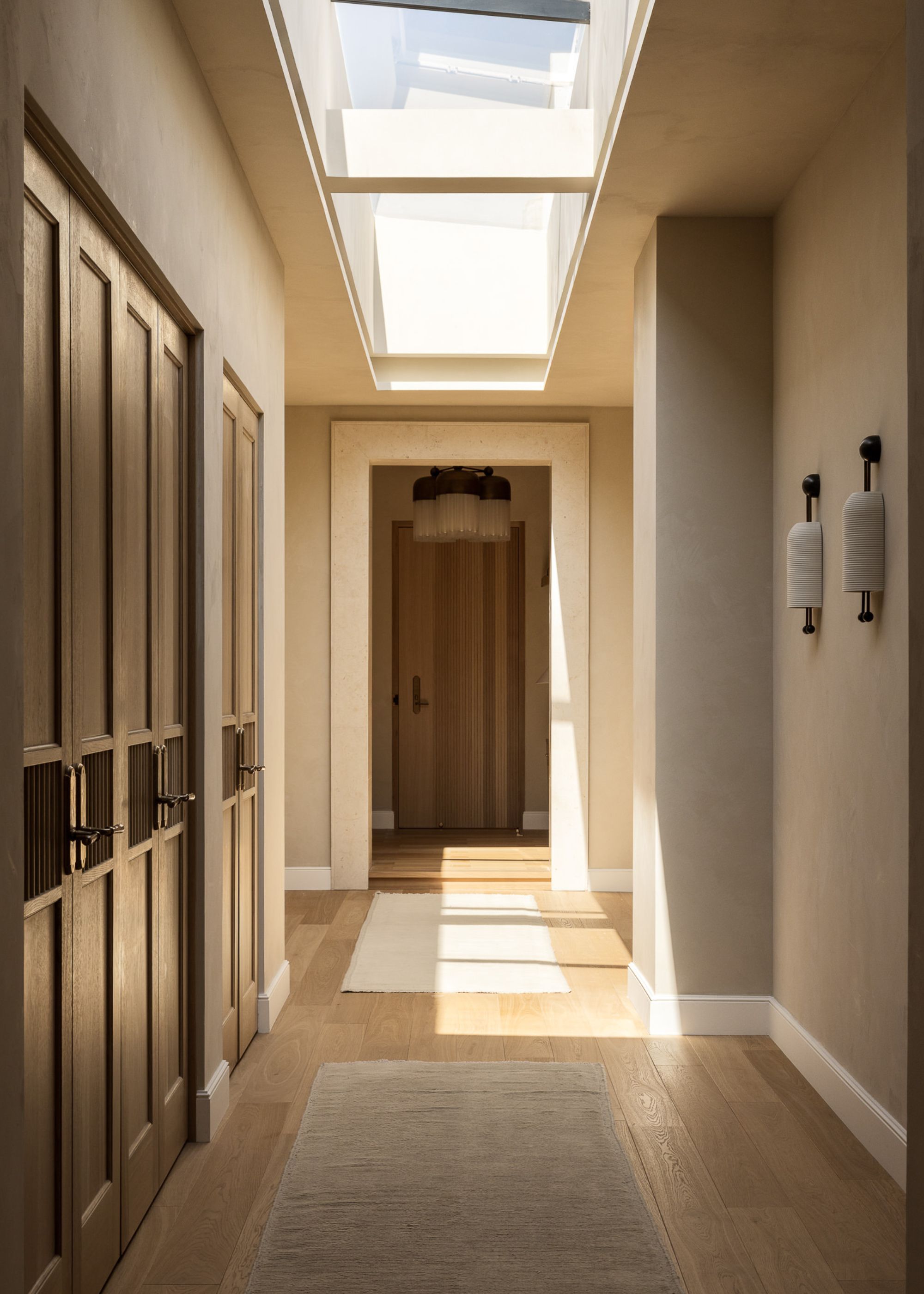 A hallway leading to the main doorway of a penthouse with skylights above and built-in cabinetry along one side of the wall with two cream rugs on the wooden flooring