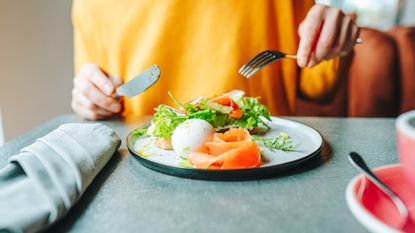 Person holding knife and fork above plae of smoked salmon, a boiled egg and salad