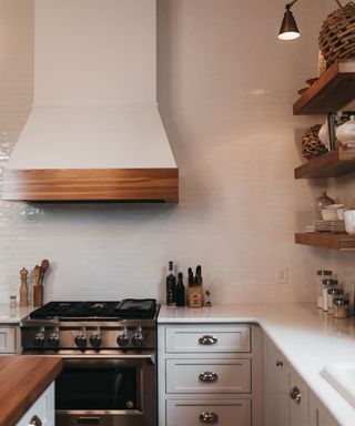 A white tiled kitchen with a large range hood, open shelves, and kitchen utensils as decor