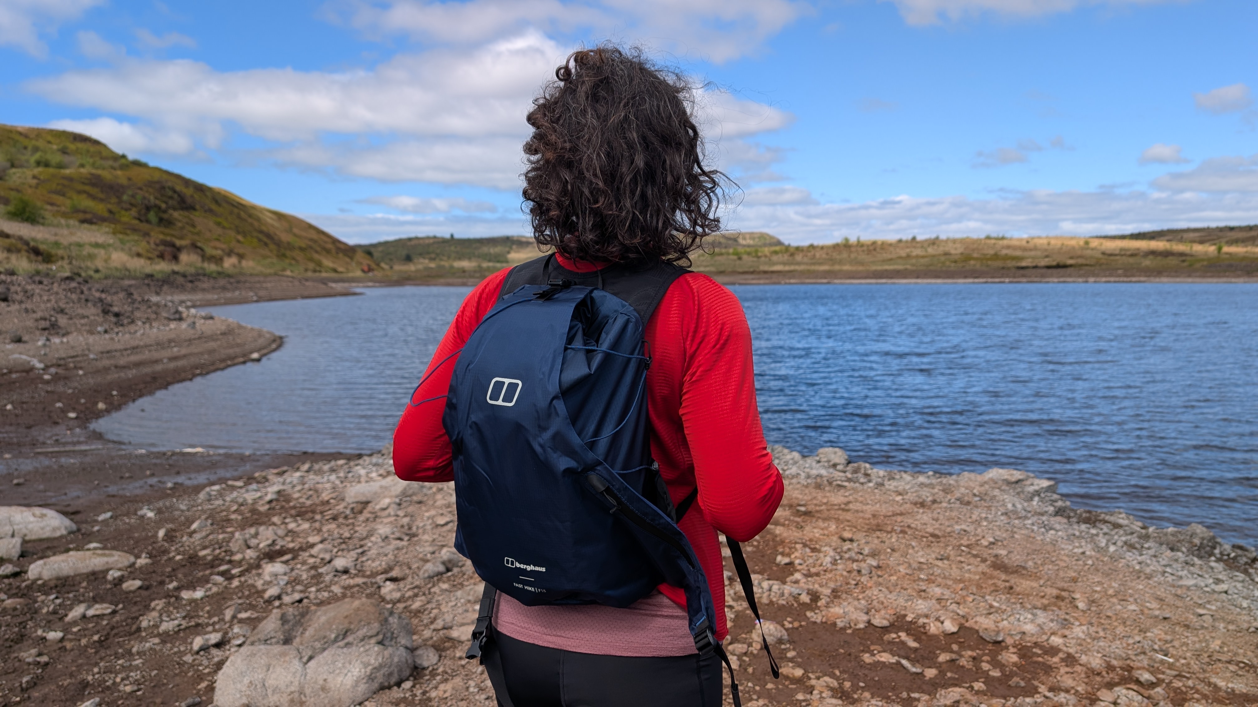 A woman hiker in a red fleece wears a blue Berghaus Fasthike F15 backpack and looks at a lake