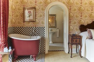 A traditional bedroom with very small pink freestanding rolltop bath in bedroom with patterned wallpaper and bed