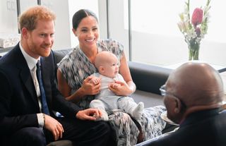 Prince Harry wears a suit with a blue tie and Meghan Markle wears a blue and white dress while holding baby Prince Archie, as they meet Desmond Tutu