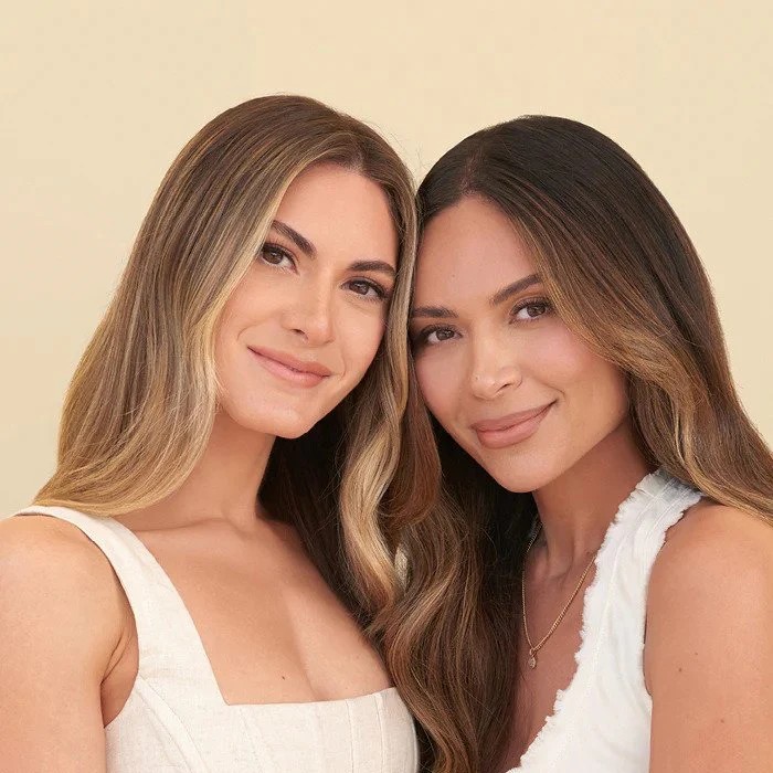 Two women wearing white tank tops smile with their heads close together.