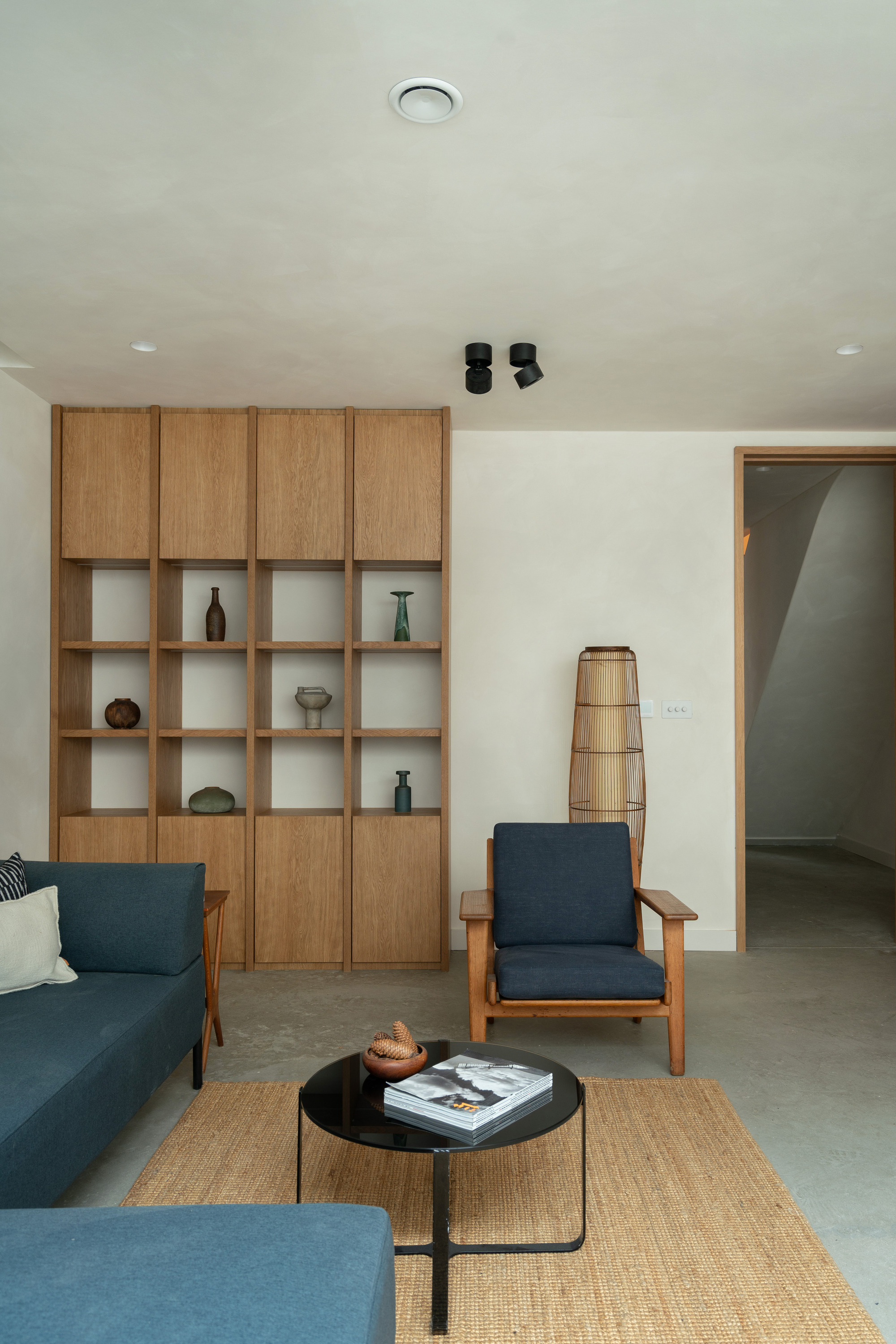modern living room with plaster walls and ceiling, polished cement floor, jute rug, blue and timber L-shaped sofa and armchair, round black coffee table with book, and built-in timber shelves styled with decor